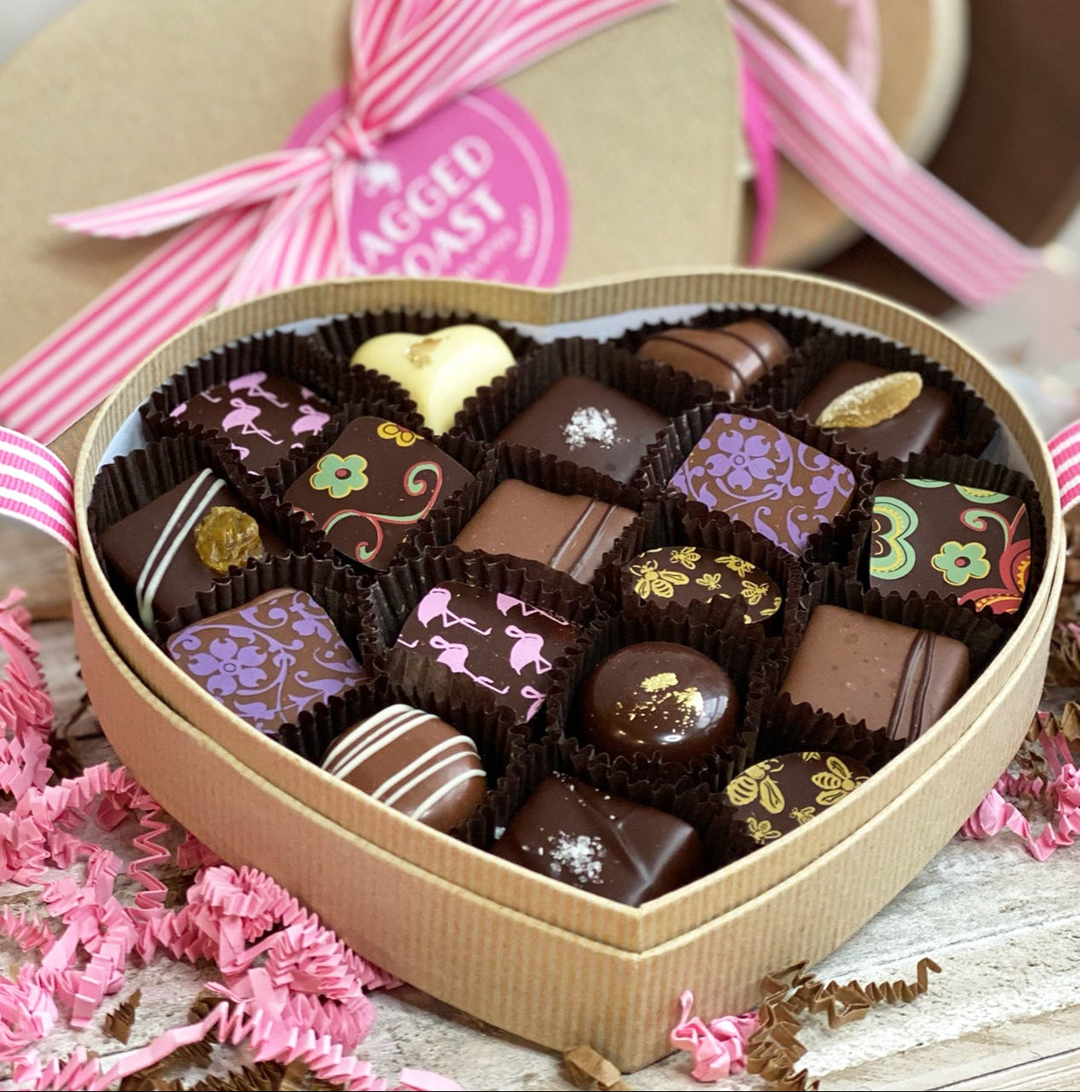 A gift basket with a large bow featuring striped ribbons in brown and pink tones. The basket contains assorted wrapped items and is filled with pink shredded paper.