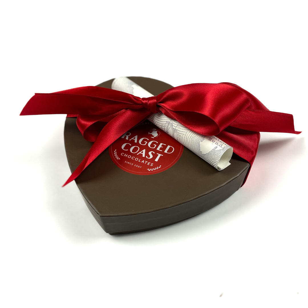 The Heart-Shaped Box - 18 Pieces from Ragged Coast Chocolates features a brown exterior, a red ribbon, and a red sticker. Inside this Grand Assortment Collection are delectable truffles, with a rolled paper elegantly tied under the bow.