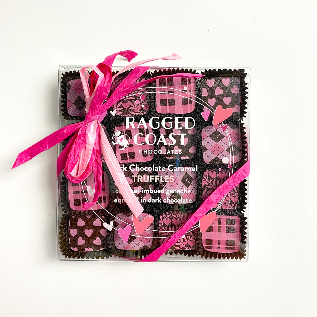 Box of Ragged Coast Chocolates with pink ribbon, adorned with heart and plaid designs on the lid. Enjoy the luxurious taste of Dark Chocolate Caramel Truffles, a treat that enhances the delightful assortment within.