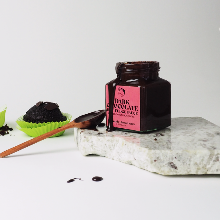 A handcrafted jar of Ragged Coast Chocolates' Dark Chocolate Hot Fudge Sauce sits on a marble slab with a wooden spoon, accompanied by chocolate desserts in green wrappers nearby. For an extra twist, try pairing it with our Salted Honey Caramel Sauce.