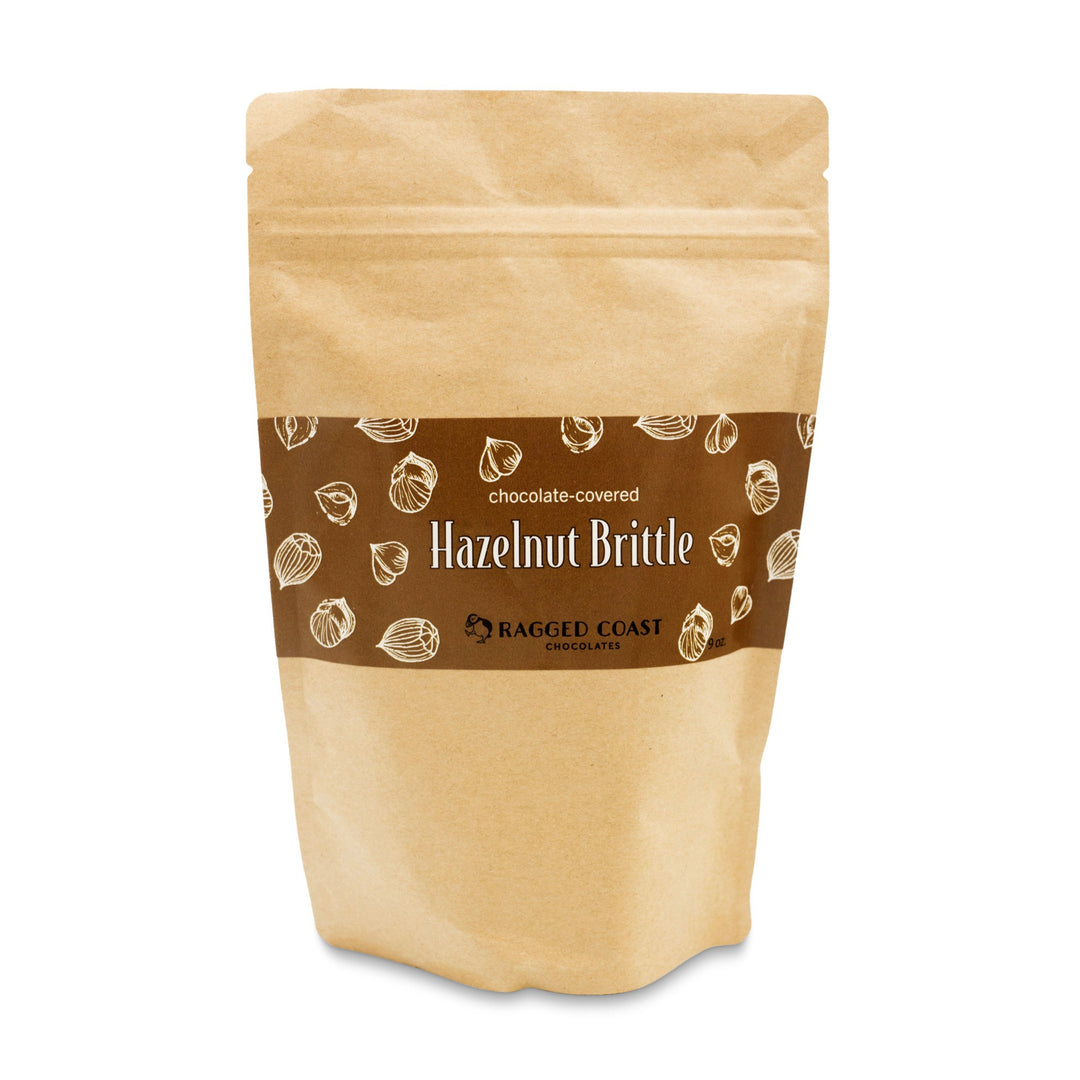 A 9-ounce brown paper bag of Milk Chocolate-Covered Hazelnut Frangelico Brittle for the Holidays by Ragged Coast Chocolates showcases white illustrations of organic hazelnuts on its label, emphasizing a delicious milk chocolate treat with hazelnut highlights.