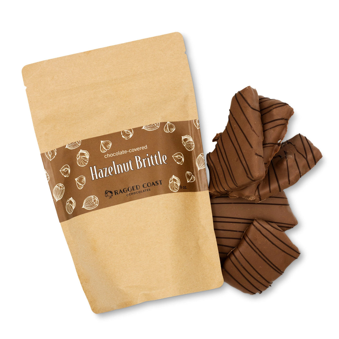 A brown paper bag labeled "Milk Chocolate-Covered Hazelnut Frangelico Brittle for the Holidays" from Ragged Coast Chocolates sits next to several pieces of chocolate-covered brittle, crafted with organic hazelnuts.