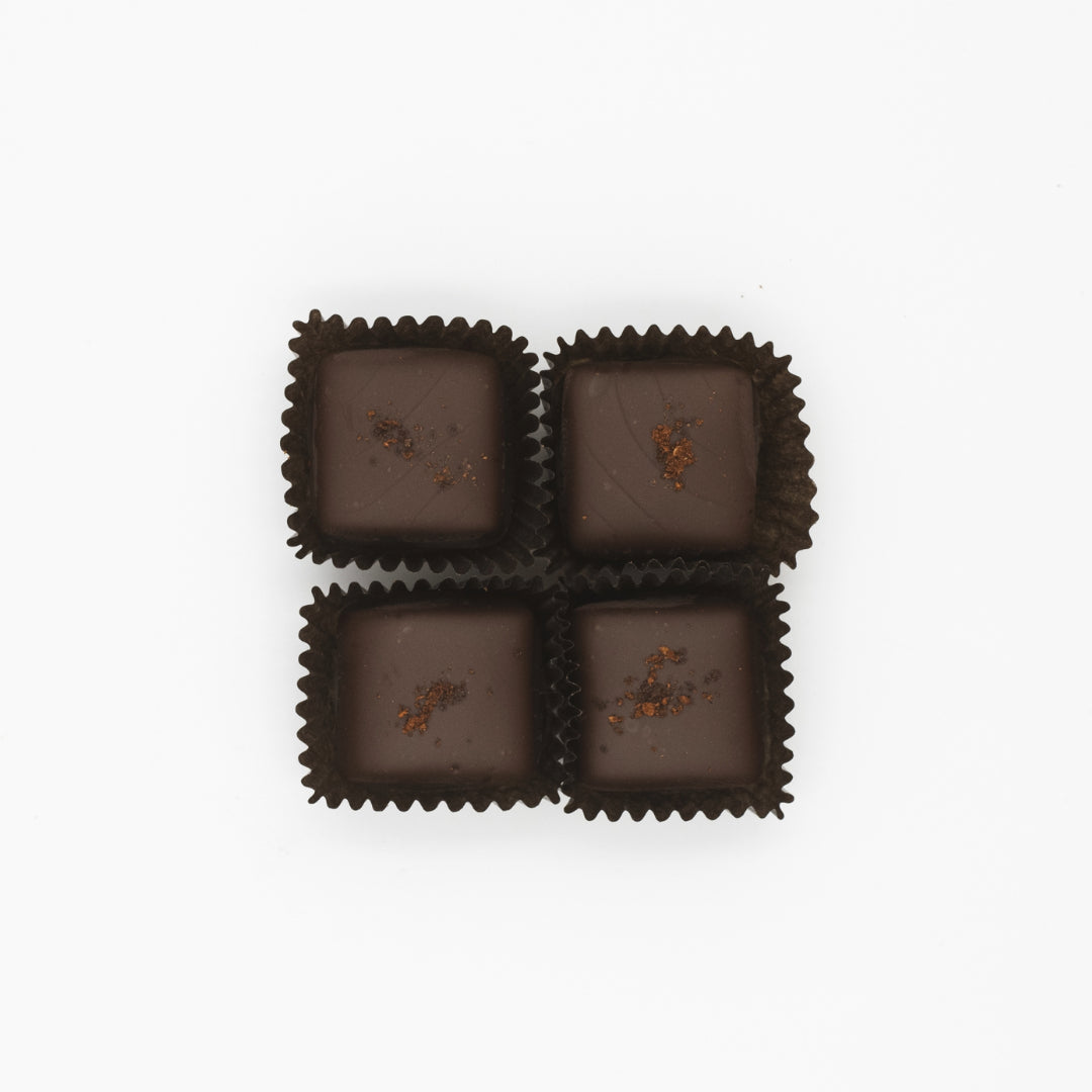 Four squares of New England Cider Caramels in Bittersweet Chocolate by Ragged Coast Chocolates, encased in sleek black wrappers and infused with cider caramel, are artfully arranged in a 2x2 grid against a pristine white background.