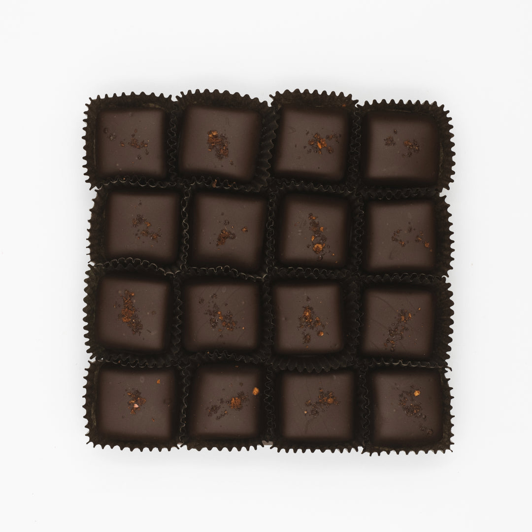 A grid of sixteen New England Cider Caramels in Bittersweet Chocolate arranged on a white background, each piece in a black paper cup and topped with a few sprinkles of apple-spiked spiced caramel. This exquisite treat is brought to you by Ragged Coast Chocolates.