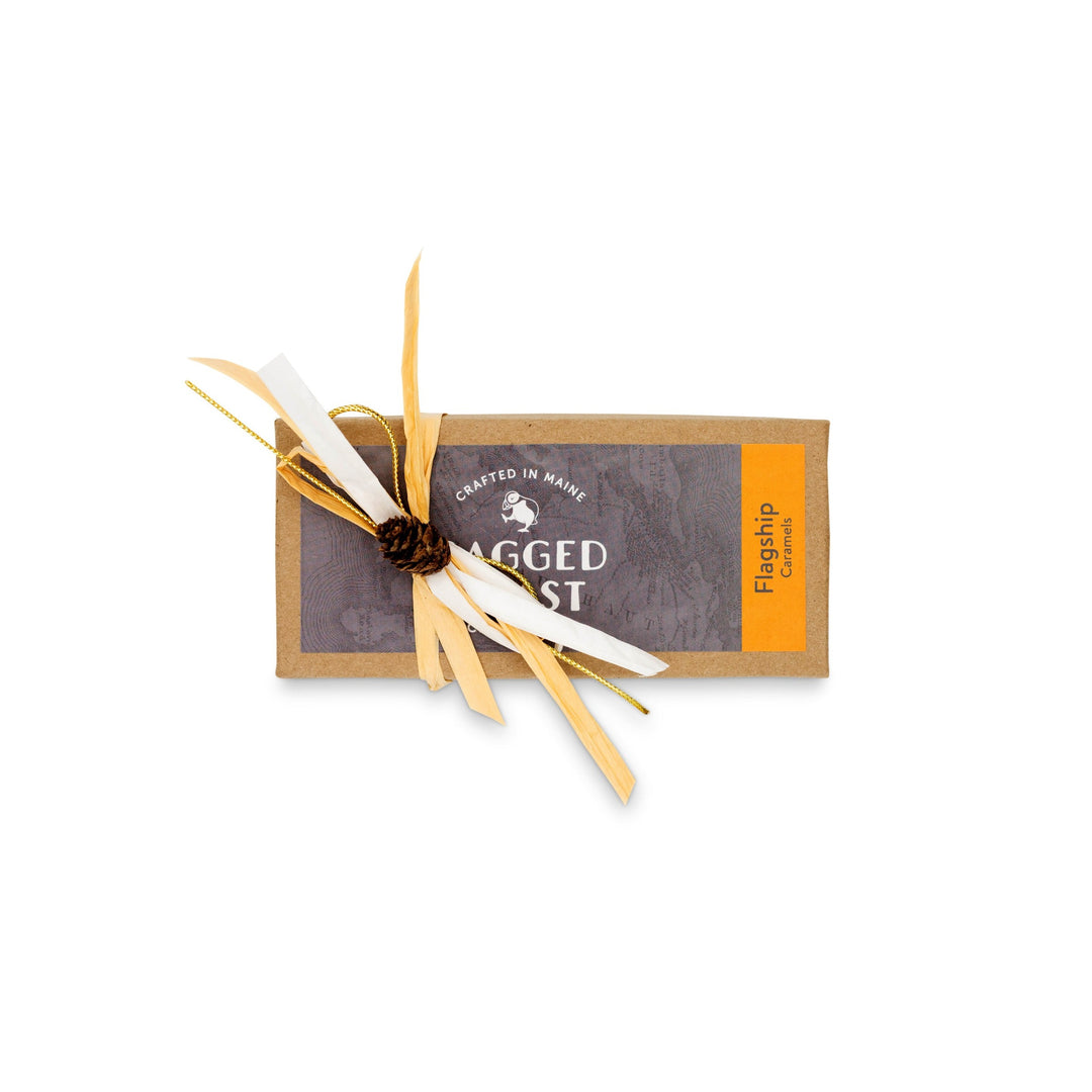 A rectangular caramel package with a grey and orange label reading "Flagship Sea Salt Caramels" and "Crafted in Maine." Infused with Maine sea salt, it's elegantly adorned with a pine cone and straw ribbons, by Ragged Coast Chocolates.