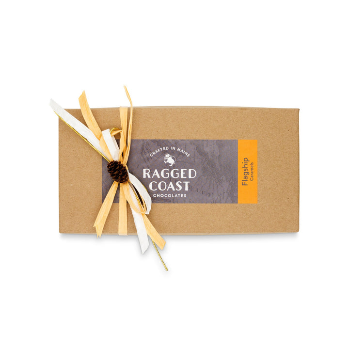 A rectangular box of Flagship Sea Salt Caramels from Ragged Coast Chocolates, adorned with a decorative bow and a small pinecone accent, unveils the indulgence of Ecuadorian dark chocolate and rich Maine sea salt.