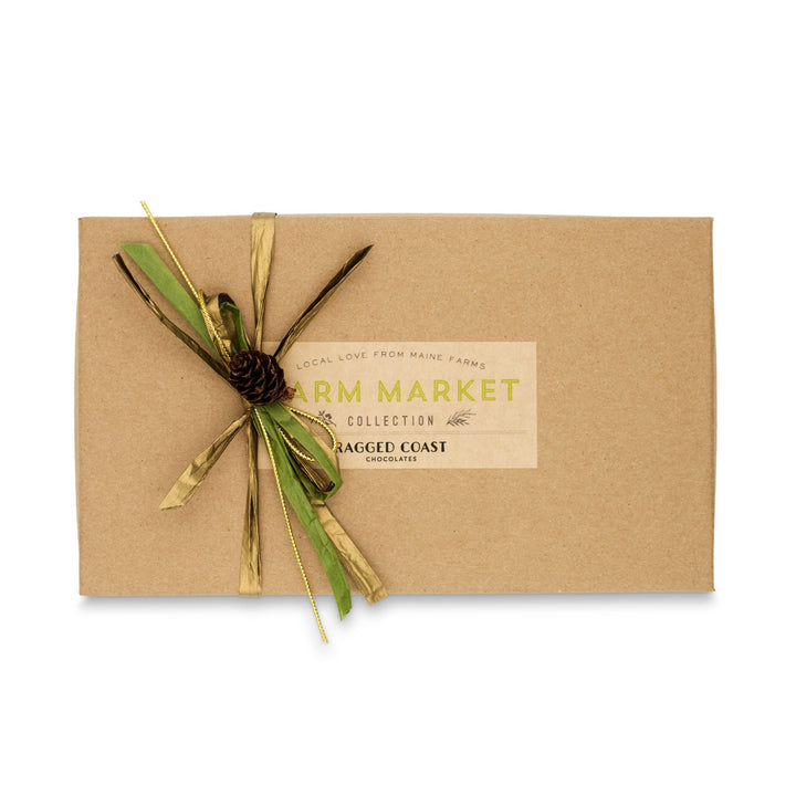 A brown gift box, decorated with a green and gold ribbon and a small pinecone, features an assortment of dark chocolate truffles. The label reads "Maine Farm Market Truffle Collection" by Ragged Coast Chocolates, celebrating Maine farm products.