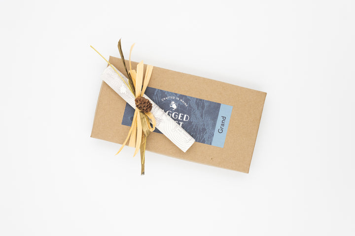 A rectangular cardboard gift box adorned with a custom ribbon and label lies elegantly on a white background, hinting at the Eighteen-Piece Chocolate Gift - Milk & Dark Chocolate Truffles by Ragged Coast Chocolates inside.