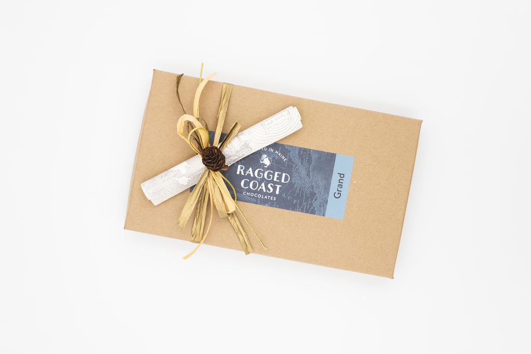 A rectangular brown box labeled "Ragged Coast Chocolates" is adorned with a natural-materials bow and contains the Twenty-Eight-Piece Chocolate Gift, featuring an exquisite assortment of milk and dark chocolate truffles.