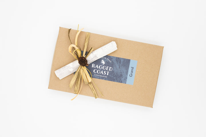 A rectangular brown box labeled "Ragged Coast Chocolates" is adorned with a natural-materials bow and contains the Twenty-Eight-Piece Chocolate Gift, featuring an exquisite assortment of milk and dark chocolate truffles.