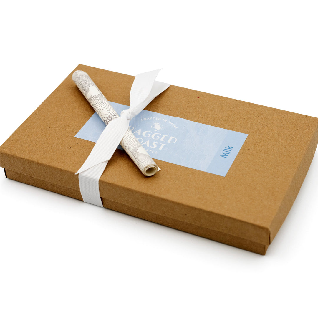 A brown rectangular box from Ragged Coast Chocolates, tied with a white grosgrain ribbon and labeled "Ragged Coast," has a rolled paper tucked beneath. Made from recyclable paperboard, it contains the delightful Milk Chocolate Truffle Assortment.