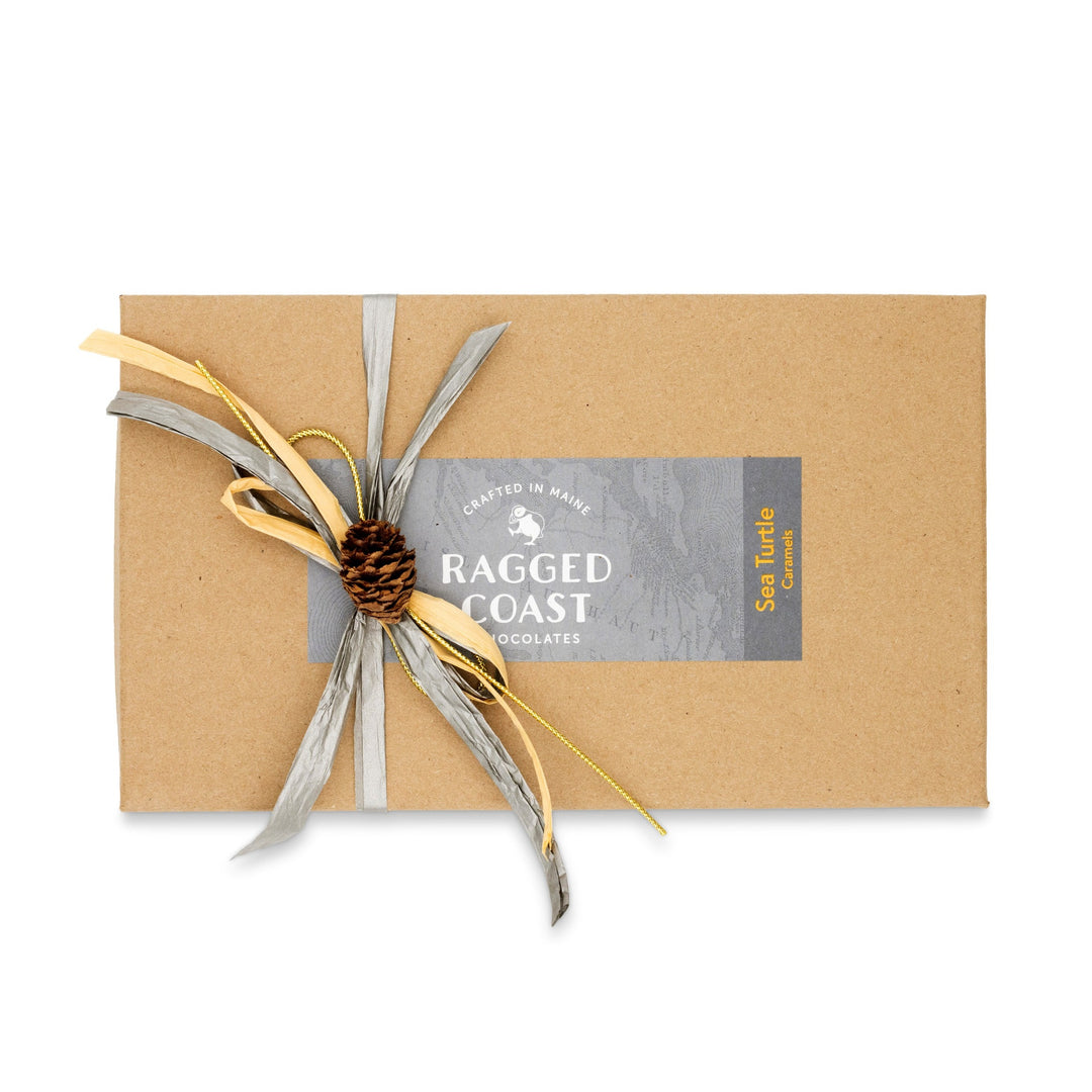 A brown gift box labeled "Ragged Coast Chocolates Sea Turtle Caramels" is tied with a decorative bow made of gray and gold ribbon, featuring a small pinecone. Inside, indulge in direct-trade Ecuadorian dark chocolate caramels that promise a rich and sustainable experience.