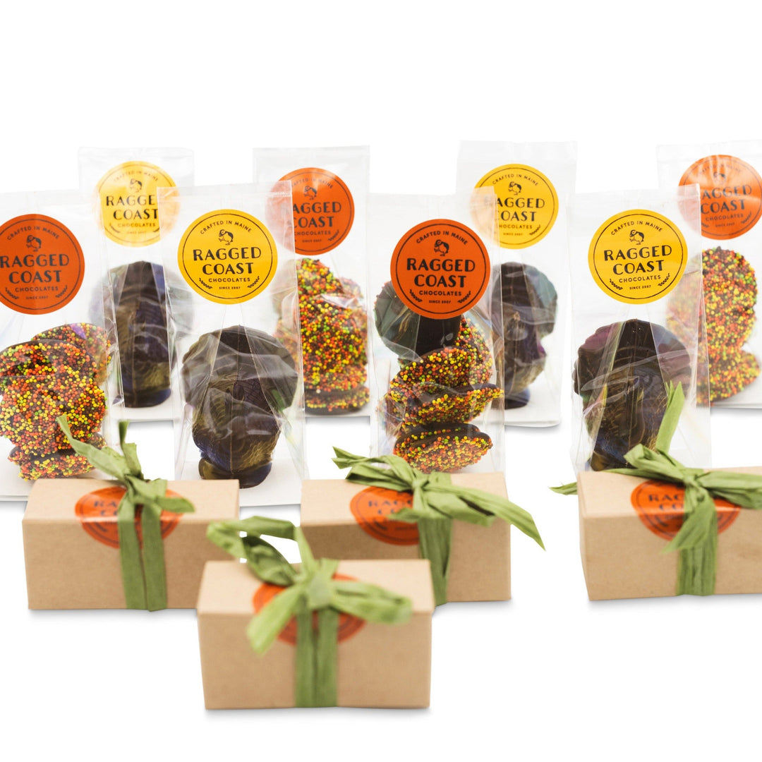 The Thanksgiving Chocolate Table Setting Bundle from Ragged Coast Chocolates features an assortment of chocolate-covered treats, including Autumn Nonpareils with colored sprinkles, along with three gift boxes tied with green ribbons.