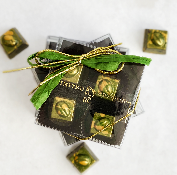 A box of Ragged Coast Chocolates' limited edition Coffee + Chocolate Irish Coffee Bonbons for Saint Patrick's Day, featuring bittersweet chocolate and pistachio toppings, tied with a green and gold ribbon, rests on a white surface.