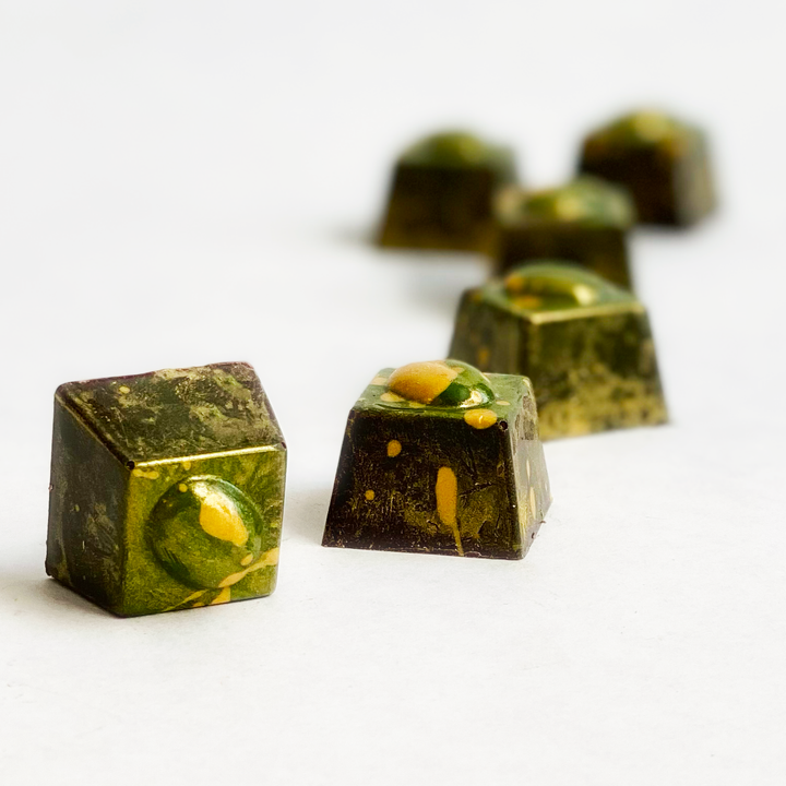 Five cube-shaped bonbons from Ragged Coast Chocolates, part of the Limited Edition Irish Coffee series for Saint Patrick's Day, with green and yellow speckles on a white surface, hide a rich whiskey ganache center.