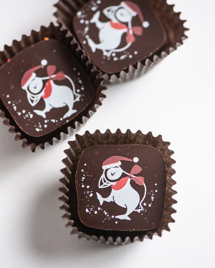 Three truffles wrapped in festive brown packaging each feature a playful penguin design, complete with a red hat and scarf. Inside these delightful Ragged Coast Chocolates' Eggnog and Bittersweet Chocolate Truffles for the Holidays is a luscious ganache, ideal for celebrating the joyous season.