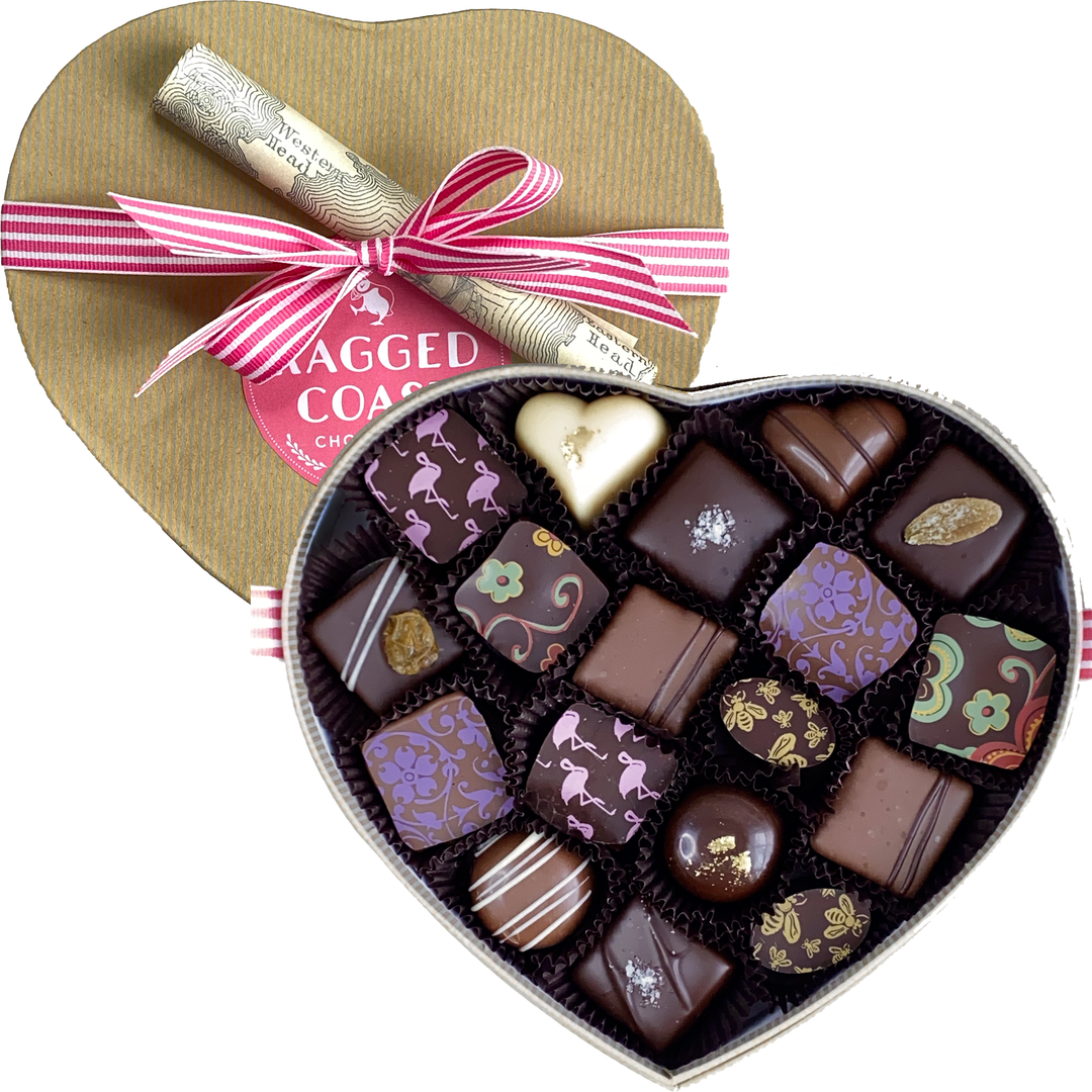 An open Heart-Shaped Box - 18 Pieces by Ragged Coast Chocolates, featuring assorted decorative chocolates and rich chocolate truffles with intricate patterns, rests beside its lid adorned with a pink and white striped ribbon.